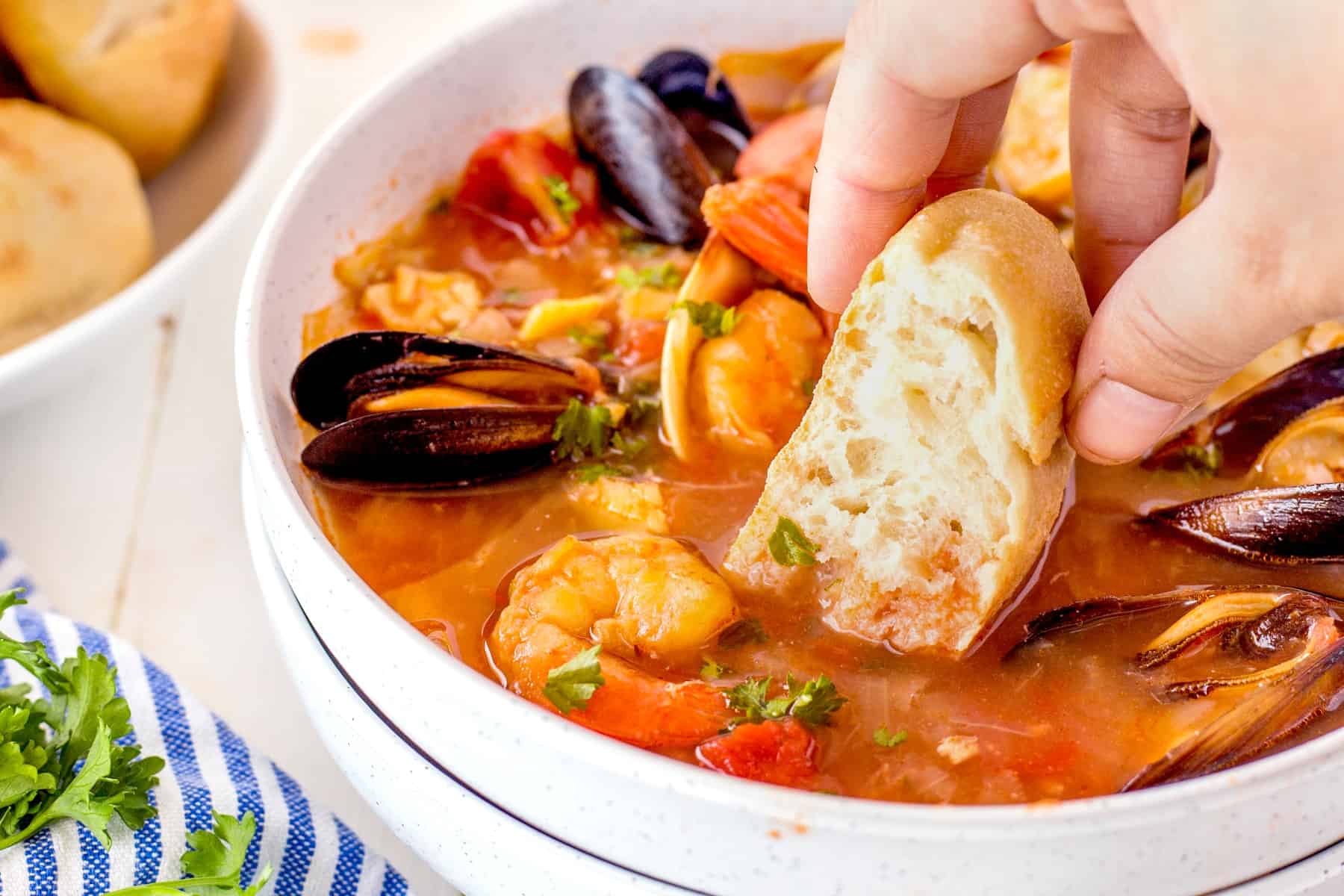 Cioppino (Fisherman's stew) is filled with shrimp, clams, mussels, white fish, and snow crab claws and simmered in a rich broth made from tomato/garlic and fish stock. Dipping bread in a bowl of Cioppino, horizontal picture.
