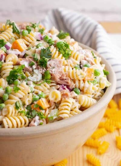 A serving bowl filled with rotini pasta, peas, tuna chunks, red onions, orange cheddar, and celery.
