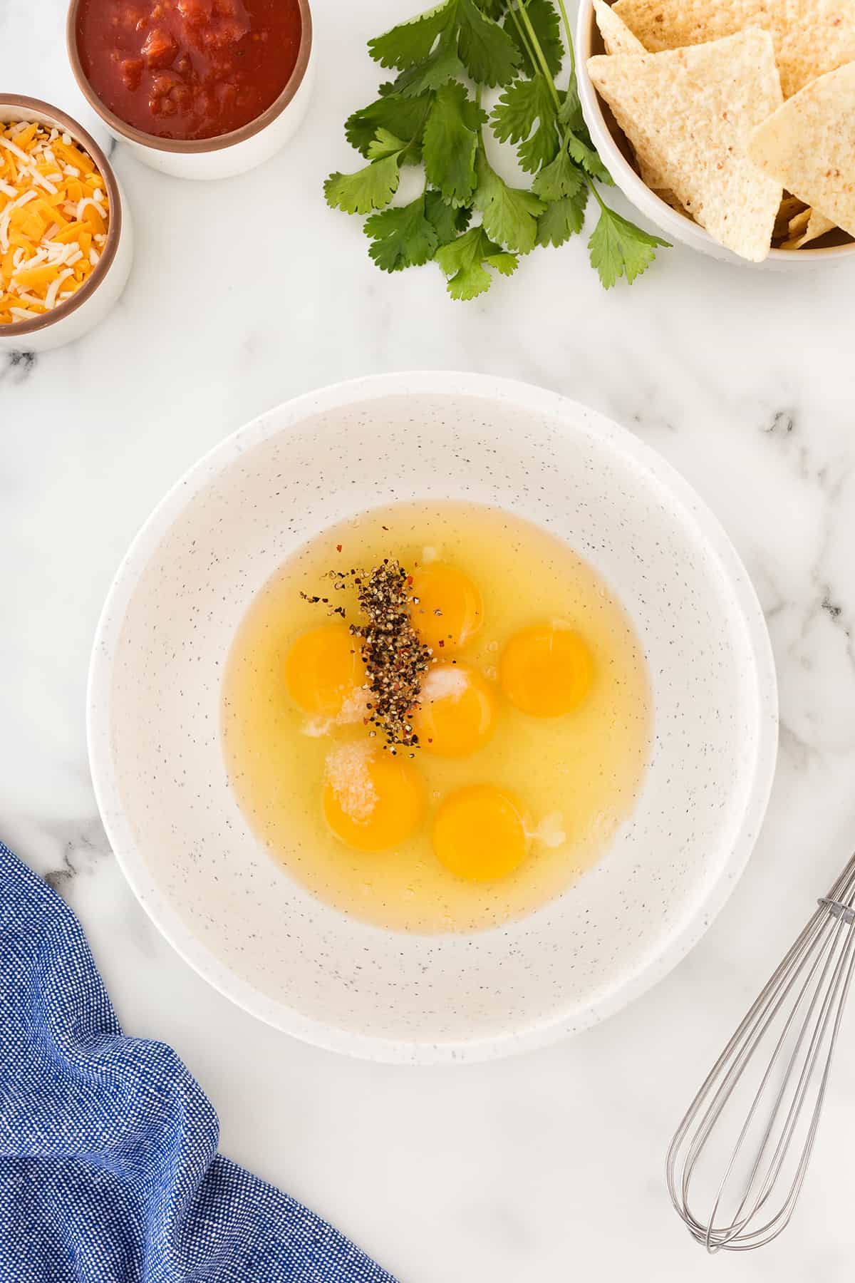 Cheesy and flavorful scrambled eggs adorn the top of crispy tortilla chips to make an unforgettable breakfast or brunch. Showing eggs in a bowl with salt and pepper. Surrounded by a whisk, cheese, salsa and chips in bowls.